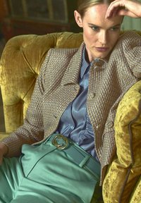 Tweed jasje in goud en blauw, satijnen lichtblauwe blouse, muntgroene broek met een ronde gouden riembuckle, zittend op een fluwelen groene stoel.