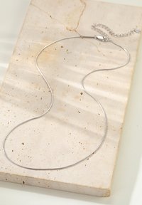 Collar de cadena de plata con un diseño suave y curvado; cuenta con un cierre y cadena ajustable en un extremo. Reposando sobre una superficie de piedra texturizada.