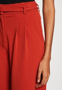 Pantalons rouges ajustés en tissu texturé, avec une ceinture étroite, des plis et une taille haute. Gros plan sur la taille et la main.
