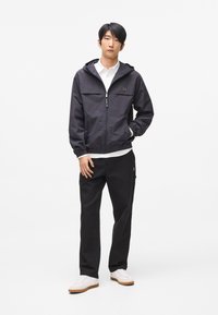 Fekete könnyű dzseki cipzárral és kapucnival, fehér ing felett viselve. Fekete nadrággal és fehér tornacipővel párosítva. Letisztult, minimalista dizájn.