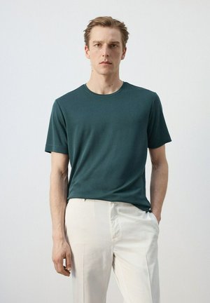 Joven con camiseta de manga corta de color verde oscuro y pantalones beige claros, de pie con una mano en el bolsillo contra un fondo liso.