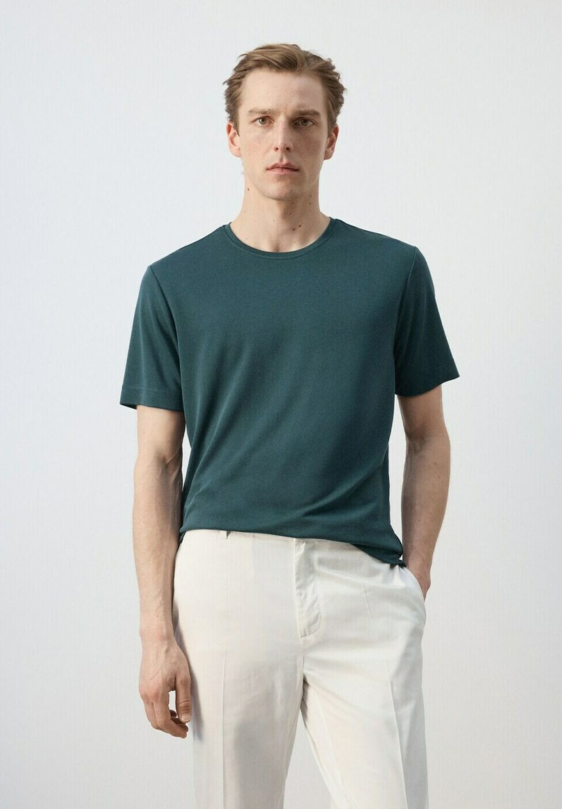 Jeune homme portant un t-shirt à manches courtes vert foncé et un pantalon beige clair, debout avec une main dans la poche devant un fond uni.