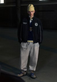 Jachetă neagră cu fermoar, cu accente albe, cămașă bleumarin dedesubt, pantaloni de trening gri și cagulă galbenă. Pantofi maro și negri cu șireturi albastre.