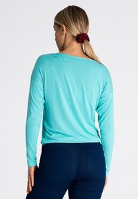 Turkusowa bluzka z długim rękawem o luźnym kroju, z dekoltem w serek i miękkiej teksturze, zestawiona z ciemnoniebieskimi rurkami.