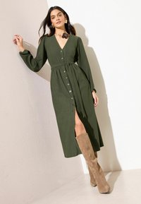 Lange groene geribbelde jurk met een V-hals, knopen aan de voorkant, een taille met riem en een zijsplit; gecombineerd met beige suède kniehoge laarzen.