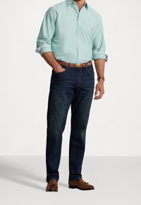Uomo in camicia verde chiaro con maniche rimboccate, jeans blu scuro, cintura marrone, orologio e mocassini in pelle marrone, in piedi con una mano in tasca.