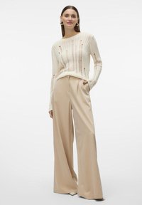 Pull en ivoire tricoté avec un côtes horizontales et de grands trous, associé à un pantalon beige à taille haute et jambes larges.