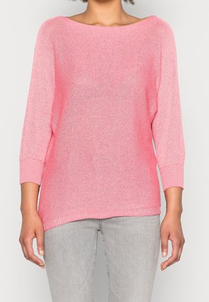 Vrouw draagt een losse roze gebreide trui met driekwartmouwen en lichtgrijze spijkerbroek, staand tegen een effen achtergrond.