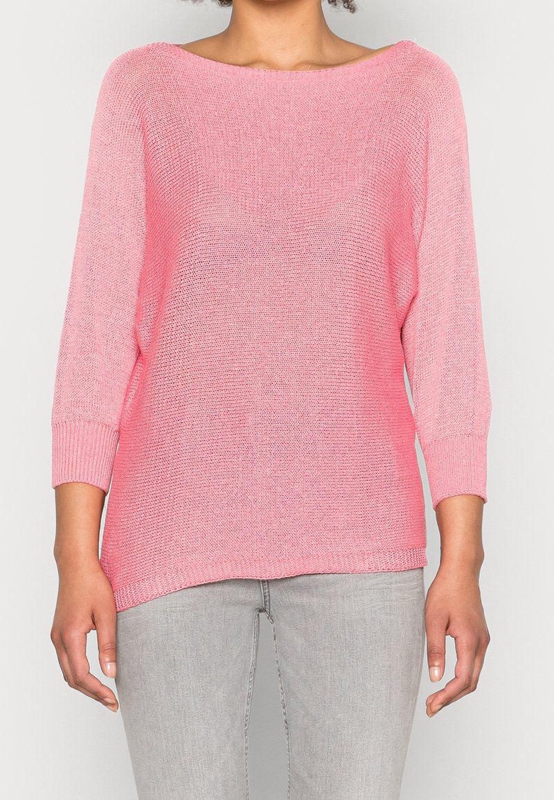 Femme portant un pull en tricot rose ample à manches trois-quarts et un jean gris clair, debout devant un fond uni.