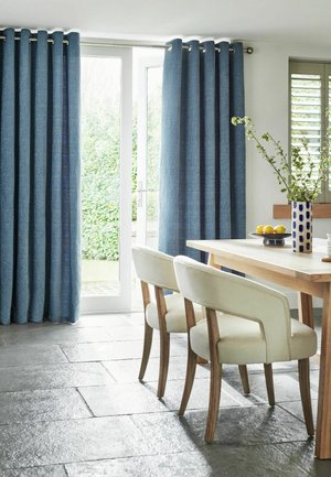Essbereich mit Holztisch, zwei cremefarbenen Stühlen, blauen Vorhängen an der Glastür und einer Vase mit grünen Zweigen auf dem Tisch.