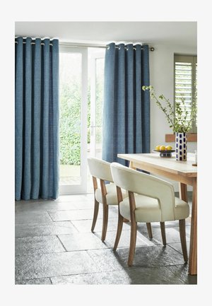 Essbereich mit Holztisch, zwei cremefarbenen Stühlen, blauen Vorhängen an der Glastür und einer Vase mit grünen Zweigen auf dem Tisch.