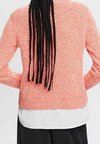 Korallinvärinen neulospusero pitkillä hihoilla ja pyöreällä pääntiellä. Päällä valkoinen paita, jonka helmasta on näkyvissä kerroksellisuus. Mustat punotut hiukset laskeutuvat alas.