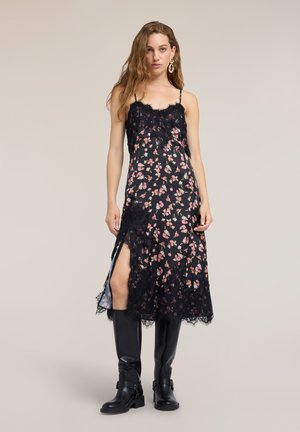 Kvinde med langt hår iført en sort blomster slip kjole med blonde detaljer og en frontslids, stylet med sorte knæhøje støvler og store øreringe.