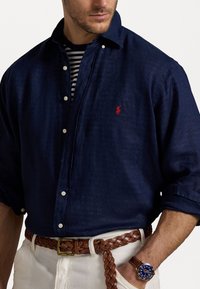 Camisa de lino azul marino con cuello abotonado y un logo rojo, combinada con un cinturón trenzado marrón y un reloj azul, llevada sobre una camisa de rayas.