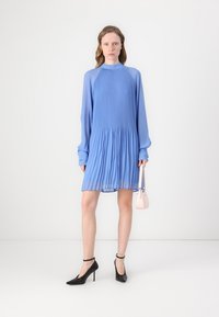 Light blue pleated dress with long sleeves and high neckline. Worn with black high-heeled shoes and a small pink handbag.