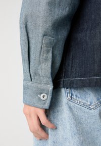 Jeansjakke med lys blå farge og en mørk blå bakpanel, knappet mansjett med metallbeslag, og en myk tekstur.