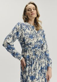 Kwiatowa sukienka w kremowym kolorze z niebieskim nadrukiem botanicznym, długie rękawy, zapięcie na guziki z przodu oraz elastyczny pas. Miękka, lekka tkanina o luźnym kroju.