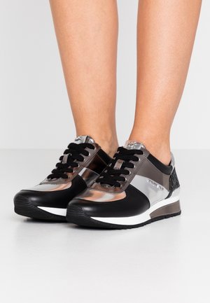 Baskets basses noires et argent avec des lacets noirs portées pieds nus sur un fond clair uni.