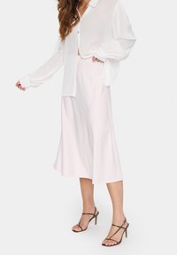 Ljusrosa satin midi-kjol med en utsvängd design, ihop med en löst sittande vit långärmad skjorta med knappar och gröna rem-sandaler.