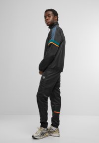 Chaqueta deportiva negra con cuello alto, que presenta rayas de colores en verde azulado y naranja. Combinada con pantalones de chándal negros y zapatillas crema.