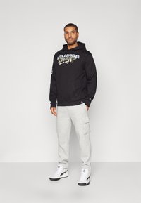 Felpa nera con logo "Los Angeles Dodgers", abbinata a pantaloni cargo grigi chiari e scarpe bianche con dettagli neri.