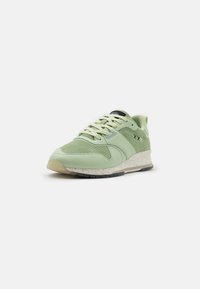 Sneaker verde chiaro con tomaia perforata, lacci, intersuola bianca maculata, suola nera e piccolo dettaglio ricamato "XXX" in bianco e nero.