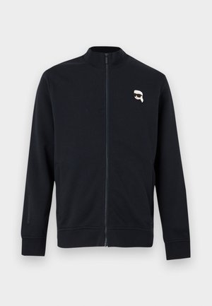 Μαύρο τζάκετ με φερμουάρ και ψηλό γιακά, μακριά μανίκια, και μικρή λευκή στυλιζαρισμένη φιγούρα με γυαλιά ηλίου στο πάνω αριστερό μέρος του στήθους.