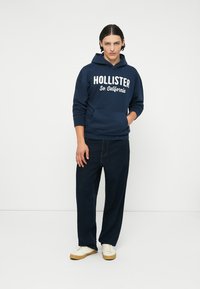Tmavě modrá mikina s kapucí s bílým potiskem "HOLLISTER So. California", klokánková kapsa, nošená s tmavými džínovými kalhotami a bílými teniskami.