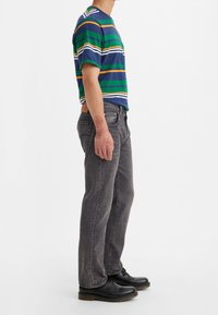 Camiseta de rayas en azul marino, verde y naranja; corte relajado. Combinada con jeans grises y zapatos negros con cordones, mostrando un diseño y textura sencillos.