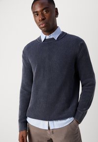 Marineblå ribbestrikket genser med rund hals, båret over en lyseblå skjorte med krage. Teksturen er myk og passformen er avslappet.