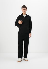 Bărbat tânăr îmbrăcat cu un polo negru cu mânecă lungă și pantaloni, ținând un clutch negru, purtând adidași albi, stând în fața unui fundal alb.