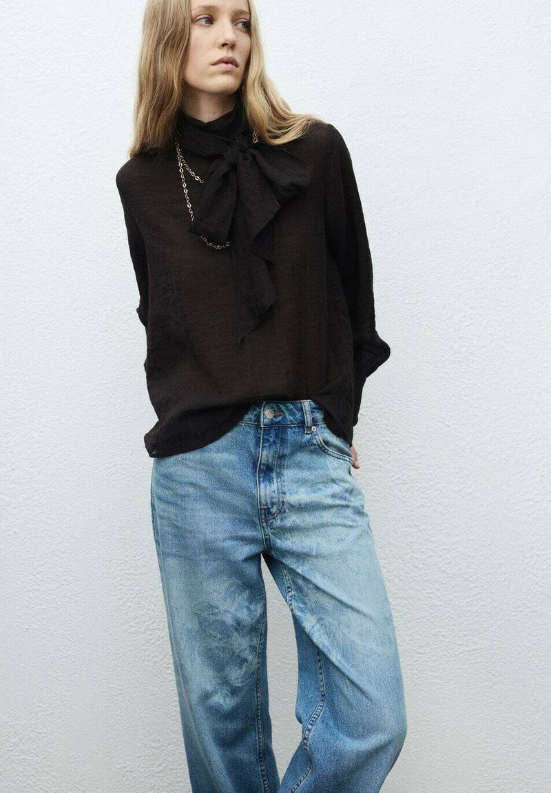 Blusa negra de manga larga con cuello de lazo, combinada con jeans de corte holgado en un tono azul claro, que presentan un desvanecimiento sutil y un diseño relajado.