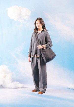 Mladá žena v sivom svetri a širokých nohaviciach, nesúca čiernu kabelku na rameno, stojaca na pozadí modrej oblohy s bielymi oblakmi.