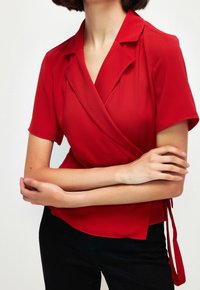 Blouse portefeuille rouge à manches courtes avec un col plié et un détail de nouage à la taille, confectionnée dans un tissu lisse. Un pantalon noir est porté en dessous.
