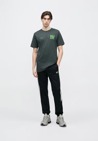 T-shirt de algodão cinzenta escura com gráficos verdes, combinada com calças de treino pretas com painéis laterais cinza claro e um pequeno logótipo. Ténis cinzentos.