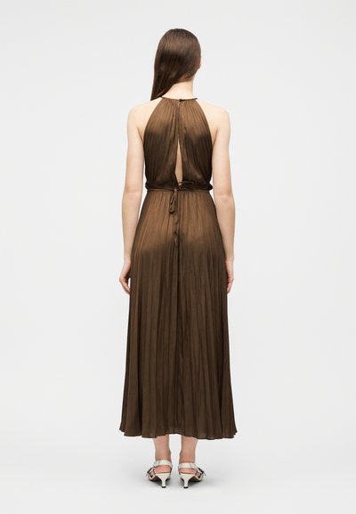 Femme portant une robe marron plissée à col licou avec une fente dans le dos et une taille nouée, associée à des sandales à talons blanches, de dos sur un fond uni.