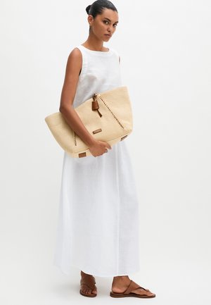 Vrouw in een witte mouwloze maxi-jurk die een grote beige geweven tas vasthoudt, bruine platte sandalen draagt, staand tegen een effen achtergrond.