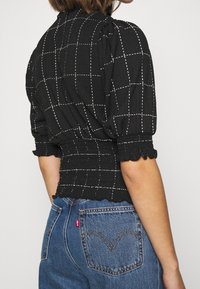 Sort tekstureret bluse med hvid rutemønster, samlet talje og flæseærmer, parret med blå denimjeans med detaljeret baglomme.