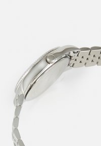 Montre-bracelet en métal argenté avec un boîtier rond, une couronne texturée et un bracelet à maillons. Le cadran de la montre est simple, sans marquages visibles.