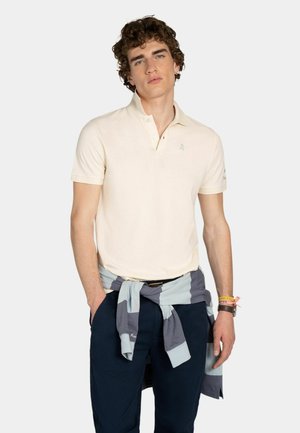 Joven con camisa tipo polo de manga corta color crema, pantalones azul marino y un suéter a rayas azul grisáceas atado alrededor de la cintura, de pie con las manos en los bolsillos.