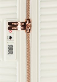 Crème-kleurige koffer met een gestructureerd oppervlak, voorzien van een dubbele koperen rits en een TSA-slot met een combinatiewiel.