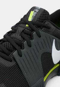 Μαύρο αθλητικό παπούτσι με πλισέ πλέγμα πάνω, γκρι συνθετικά στοιχεία, πράσινες λεπτομέρειες και μαύρες κορδόνες. Διαθέτει μια εξέχουσα λευκή λογότυπο.