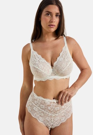 Femme portant un soutien-gorge en dentelle florale blanche et un slip taille haute en dentelle, debout devant un fond clair uni.
