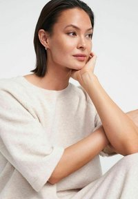 Lys beige oversized sweater med korte ærmer. Blødt, tekstureret stof. Model siddende, med hagen hvilende på hånden, med glat hud og kort hår.