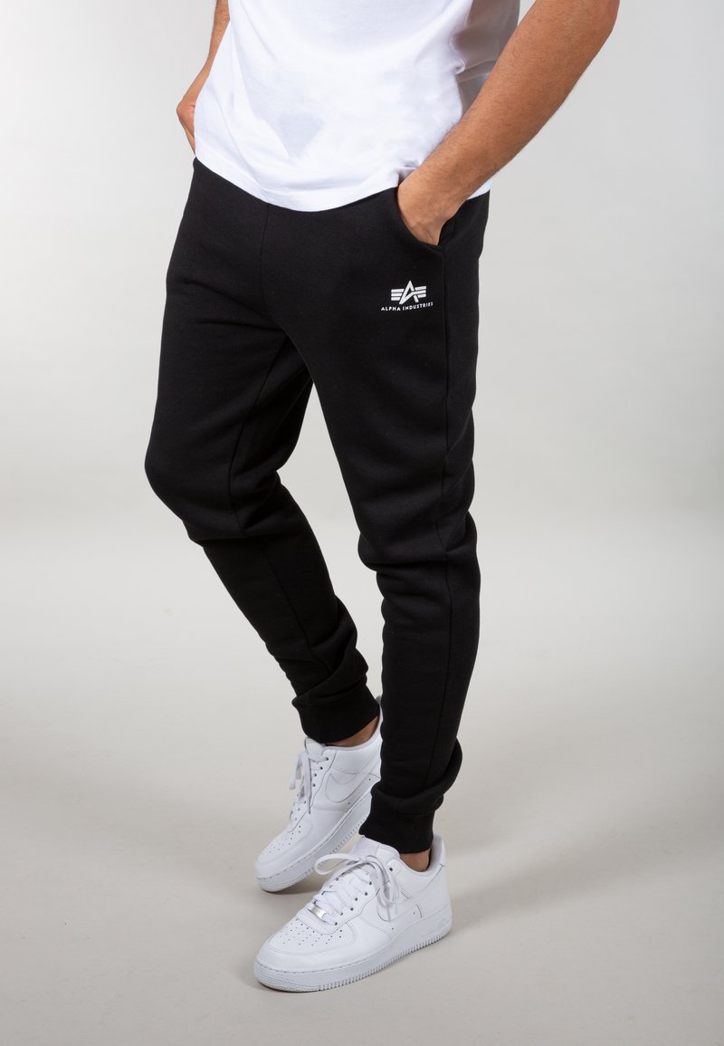 Mand iført sorte Alpha Industries joggers og hvide sneakers, stående med hænderne i lommen mod en lys baggrund.