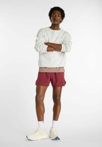 Felpa grigio chiaro con vestibilità morbida e polsini a coste, indossata sopra una camicia color taupe. Pantaloncini color marrone borgogna, sneakers crema con dettagli blu navy.