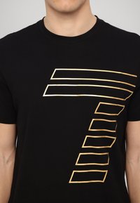 Czarny bawełniany t-shirt z krótkimi rękawami, na piersi z złotym liniowym wzorem w kształcie cyfry siedem.