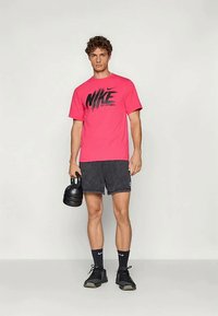 Rozā Nike T-kreklā ar melnu logotipu, savienots ar tumšiem šortiem. Modelis tur melnu kettlebellu un valkā melnas sporta kurpes un zeķes.