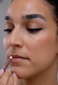 Konturówka do ust trzymana blisko warg, półmatowe wykończenie, jasnobrązowy odcień. Gładka tekstura skóry, wyraźnie podkreślona kredka do oczu, naturalny kolor ust.