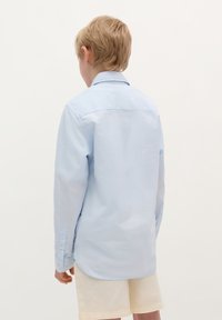 Camicia a maniche lunghe azzurro chiaro con colletto, polsini abbottonati e orlo curvo. Il tessuto appare liscio e leggero. Indossata con pantaloni corti beige.
